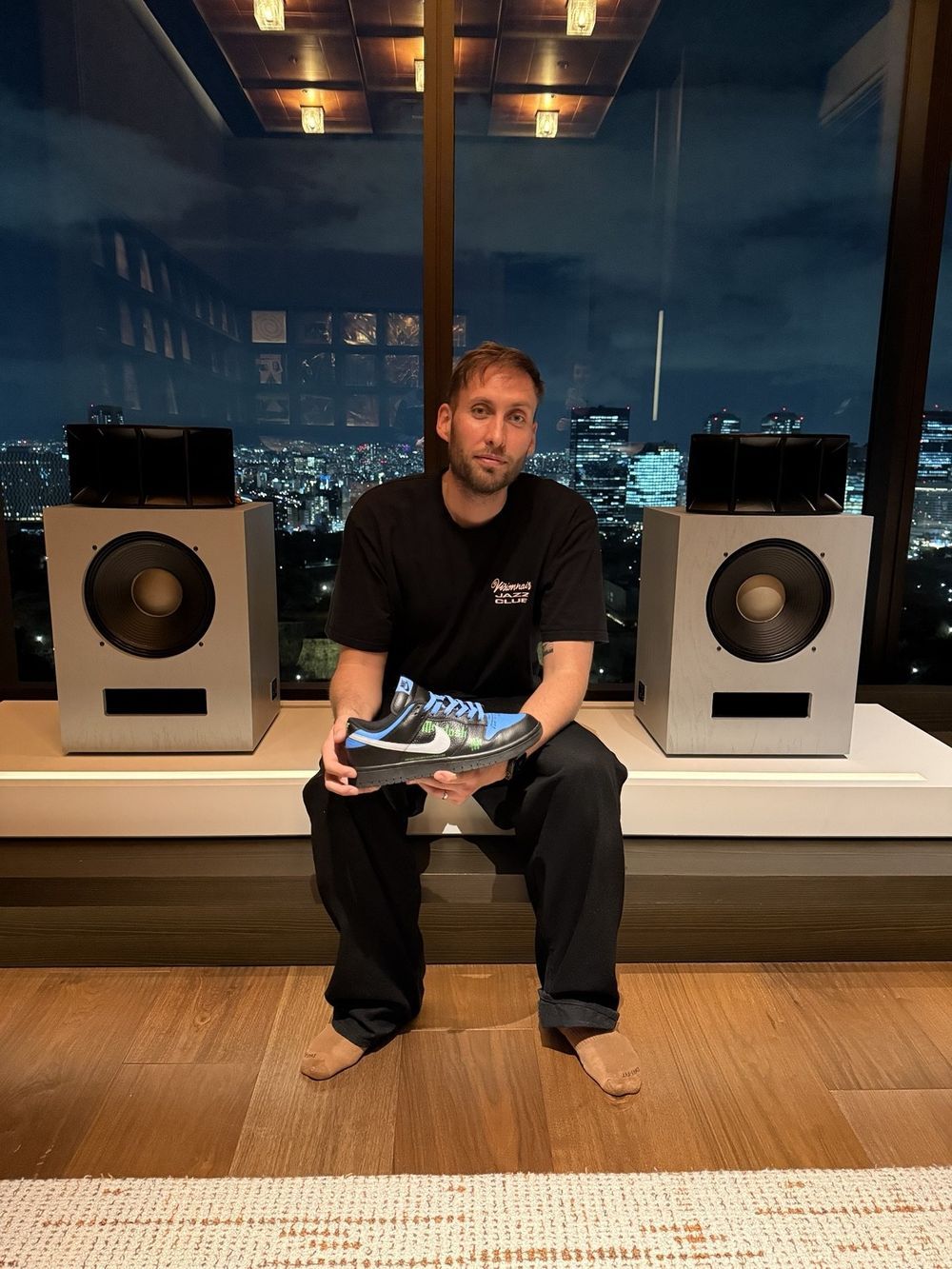Hugo Lahutte assis devant deux enceintes Klipsch dans une salle d'écoute d'Osaka, la paire McIntosh à la main, la ville éclairée derrière la baie vitrée.